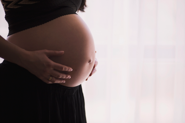 Eine schwangere Frau in einem weißen Kleid steht vor einem offenen Fenster, die Hände auf dem Bauch, mit einem friedlichen Ausdruck, beleuchtet von natürlichem Licht.