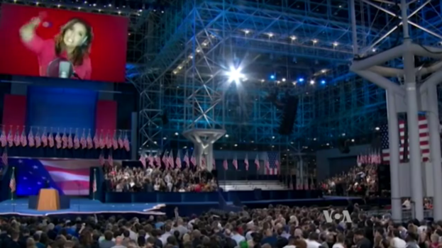 Eine große Menschenmenge sitzt vor einer Bühne mit einem Rednerpult, Mikrofon, Flaggen, einem Bildschirm mit einer Frau und Lichtausrüstung und wartet auf eine Präsidentschaftsdebatte.