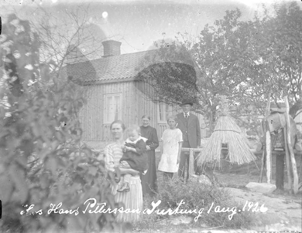 Ein altes Schwarz-Weiß-Foto einer Familie, die vor einem Haus steht, mit einer Frau, die ein Baby hält, umgeben von anderen Familienmitgliedern, Bäumen und einer Hütte im Hintergrund, und Text unten.