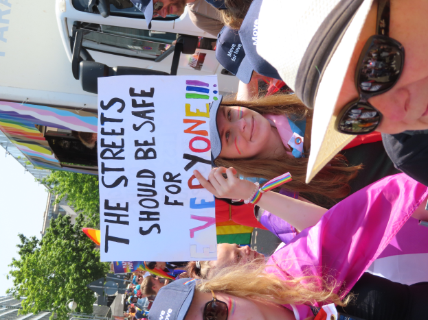 Eine Gruppe von Menschen steht vor einem Truck und hält ein Schild hoch, auf dem steht "Die Straßen sollten sicher sein". Einige tragen Mützen und Schutzbrillen, andere halten Schilder, vor einem Hintergrund aus Bäumen, Gebäuden und einem klaren blauen Himmel.