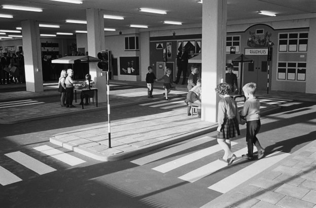 Schwarzes und weißes Foto von Kindern, die eine Straße an einer Ampel überqueren, mit Fußgängern in der Nähe, einem Verkehrssignalmast im Vordergrund und strukturellen Pfeilern und Deckenleuchten im Hintergrund.