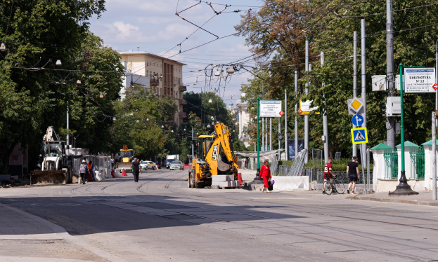 Eine Stadtstraße mit einer Baustelle, Fahrzeugen, Fußgängern, einem Radfahrer, Verkehrskegeln, Pfosten, Hinweisschildern, Strommasten mit Kabeln, Bäumen, Gebäuden mit Fenstern und einem bewölkten Himmel.