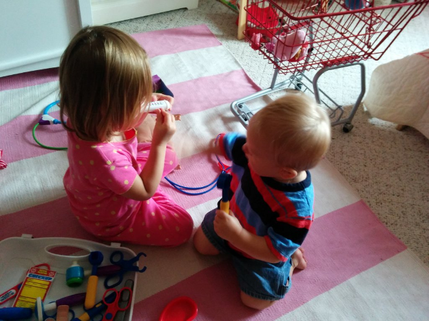 Kinder sitzen in der Mitte, ein Wagen auf der rechten Seite mit Gegenständen darauf, ein weißes Objekt in der Nähe, ein Tablett unten links, eine Matte auf dem Boden mit einem Draht und einem roten Objekt darauf.
