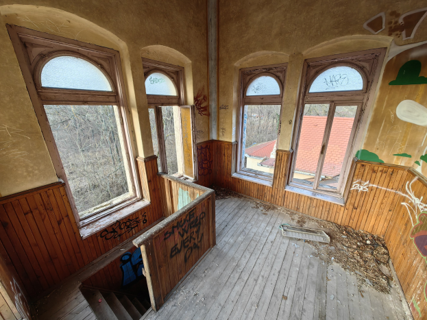 Verlassenes Gebäude mit graffiti-bedeckten Wänden und Fenstern, verstreuter Trümmer, einer hölzernen Treppe und einem sichtbaren Boden, umgeben von Bäumen und einem fernen Haus.