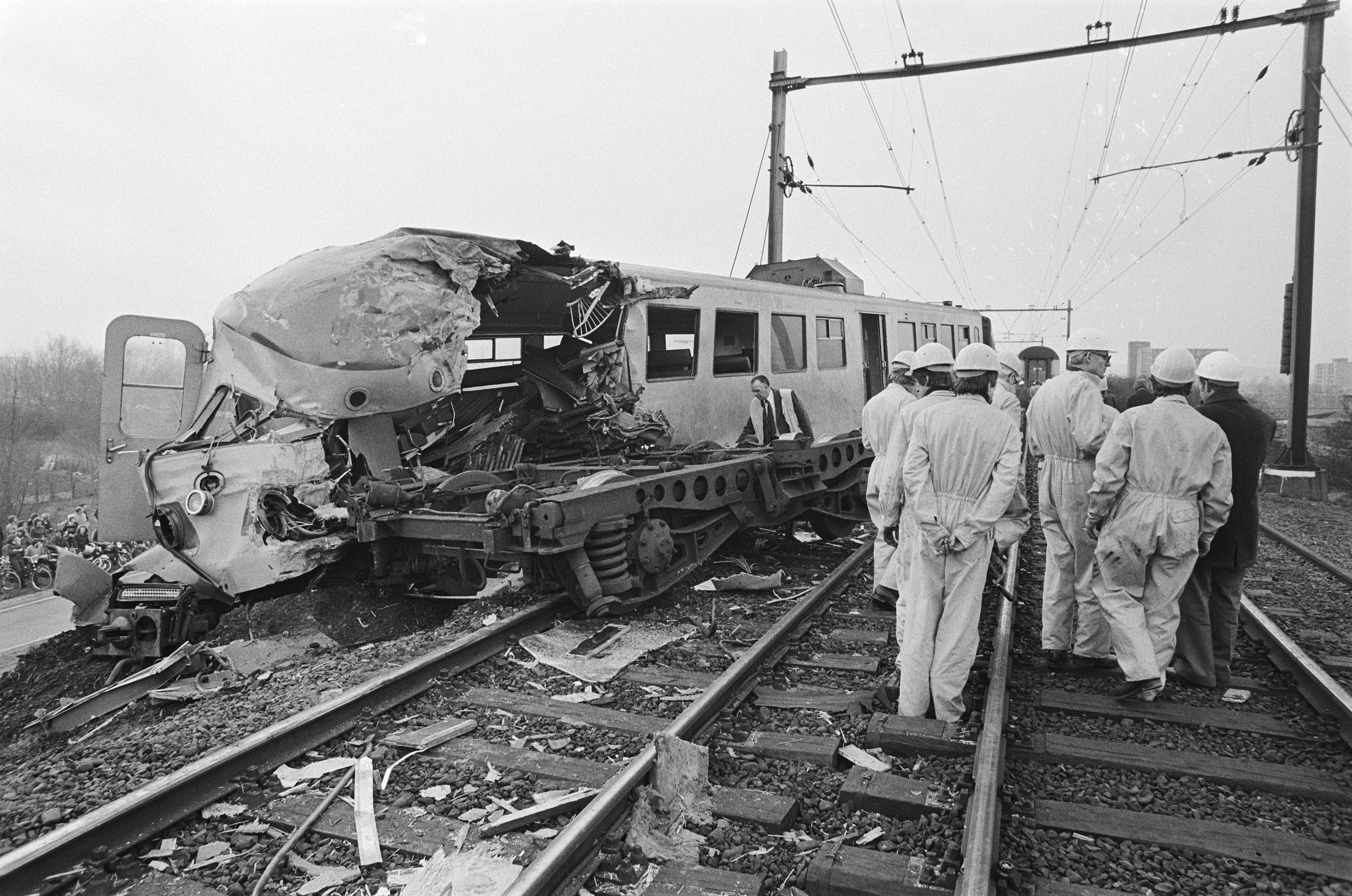 Ein Schwarz-Weiß-Bild einer Zugkollision mit Menschen in Helmen drumherum, Strommasten, Bäumen, Gebäuden und einem klaren Himmel im Hintergrund.
