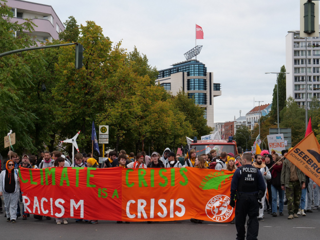 Eine Gruppe von Menschen marschiert auf einer Straße, hält ein Banner mit der Aufschrift "Klima-Krise ist eine Krise", mit Bäumen, Laternenpfählen und Schildern an der Straße, einem geparkten Fahrzeug in der Nähe, Gebäuden im Hintergrund und einem klaren blauen Himmel.