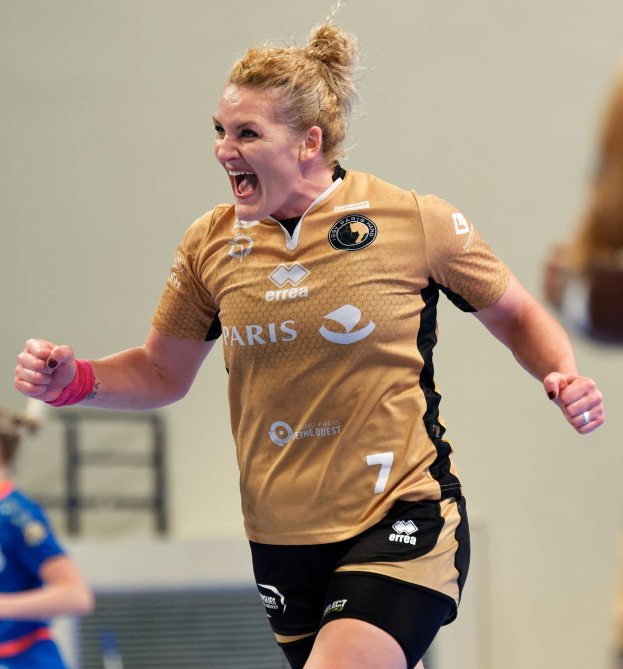 Eine Frau in braunem T-Shirt und schwarzen Shorts rennt auf einem Futsal-Platz und feiert mit triumphierendem Gesichtsausdruck, während zwei andere Frauen im Hintergrund unscharf sind.