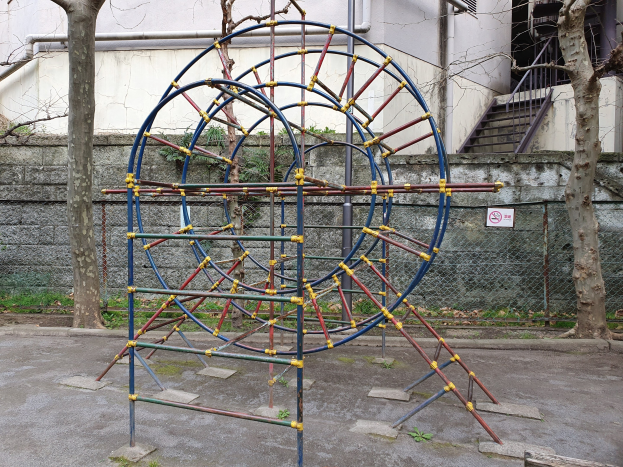 Spielplatz mit großem Metallklettergerüst, umgeben von Bäumen und einer Maschendrahtzaun, mit Treppen mit Geländern, die zu einem Gebäude mit Fenstern und Rohren führen, einem Schild an einem Pfahl und Grasboden.