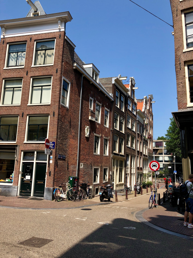 Eine Straße in Amsterdam, Niederlande, mit Gebäuden auf beiden Seiten, Menschen auf dem Gehweg, parkenden Fahrrädern, Schildern, Bäumen und einem klaren blauen Himmel.