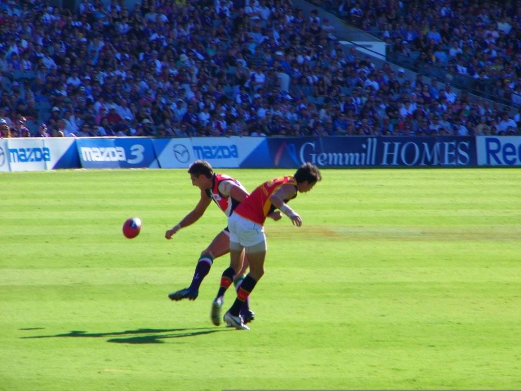 Zwei Personen in rot-weißer Kleidung stehen mit einem Ball in der Luft, während Banner und eine Zuschauermenge im Stadionhintergrund zu sehen sind.