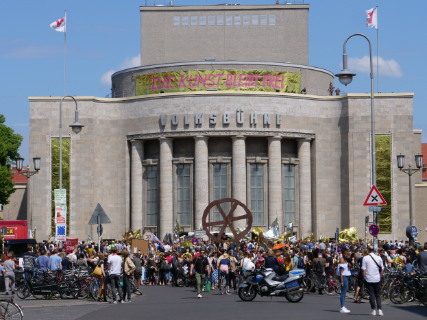 Eine große Gruppe von Menschen versammelte sich vor einem Gebäude mit Säulen und Text, umgeben von Straßenmasten, Lichtern, Schildern, Motorradfahrern, Kraftfahrzeugen, Bäumen und einem bewölktem Himmel während einer Demonstration in Berlin, Deutschland.