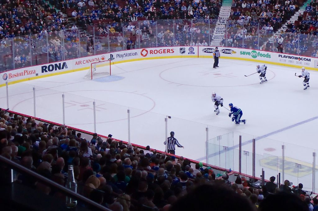 Vier Männer spielen Hockey auf einem Eisstadion, mit zwei in der Nähe des Tors und des Zauns stehenden Männern, Zuschauern auf Stühlen im Vorder- und Hintergrund sowie einer Treppe im Hintergrund.