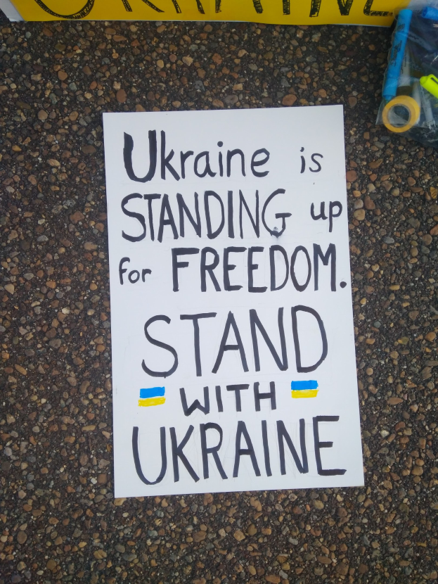 Weißer Zettel mit 'Ukraine steht für Freiheit, steht mit Ukraine' vor einem gelben Banner, rechts daneben eine Plastikhülle mit verschiedenen Gegenständen.