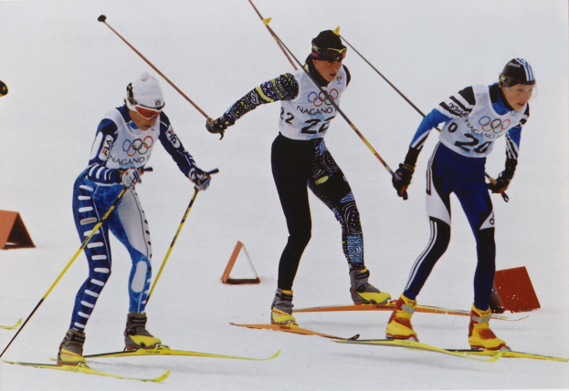 Drei Skifahrer sausen eine schneebedeckte Piste hinunter, jeder hält Ski-Stöcke und trägt bunte Ski-Anzüge und Helme, während der Schnee in der Sonne glitzert.