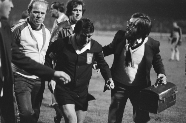 Schwarzes und weißes Bild einer Gruppe von Männern, die auf einem Fußballfeld gehen, einer hält eine Aktentasche und sieht besorgt aus, mit einem Fußballspieler in der Mitte.