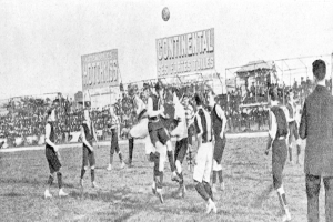 Ein Schwarz-Weiß-Bild einer Gruppe von Männern, die auf einem Feld Fußball spielen, mit einem Ball in der Luft und Zuschauern im Hintergrund, mit Tafeln, die Text über das Spiel anzeigen.