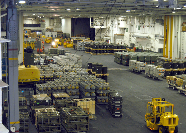 Ein großes Lagerhaus voller Numerous Trucks, Container, Menschen, Lichter, Stangen und andere Gegenstände, mit einem prominenten Rad.