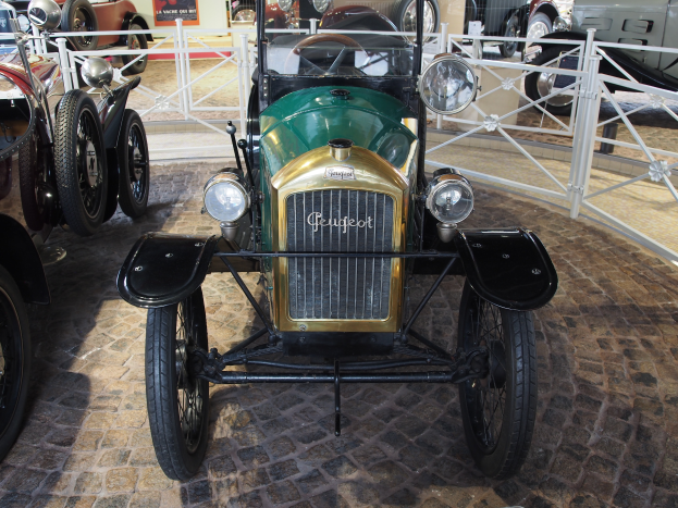 Ein Oldtimer in einem Museum, umgeben von anderen Autos und einem Geländer, mit Informationsschildern im Hintergrund.
