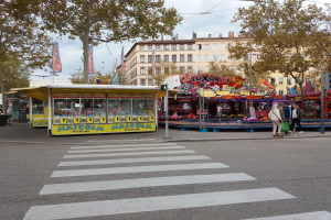 Eine belebte Straße mit Zebrastreifen und einem Karussell, mit Menschen, Stönden, Bäumen, Gebäuden, Bannern, Laternenmästen und einer Ampel unter einem bewölktem Himmel.