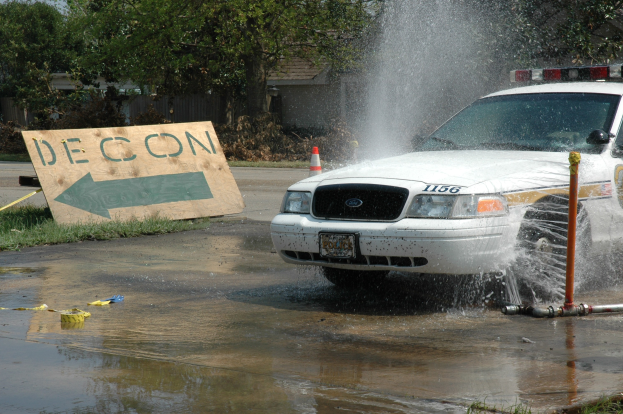 Polizeifahrzeug fährt durch eine Pfütze mit verstreuten Gegenständen, darunter eine Tafel mit Text und ein Verkehrskegel, vor einem Hintergrund aus Bäumen, Gebäuden und einem klaren blauen Himmel.