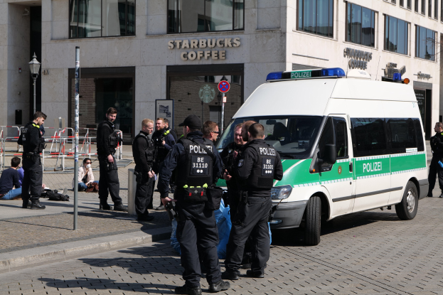 Polizeibeamte vor einem Starbucks-Café mit einem Van auf der rechten Seite und Menschen auf der linken Seite, vor einer Gebäudefassade, einem Schild, einer Straßenlaterne und einem Zaun.