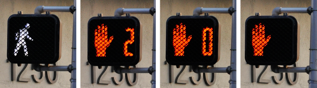 Eine Ampel mit drei Signalen an einem Gebäude angebracht, zeigt rote, gelbe und grüne Lichter vor einem Wandhintergrund.
