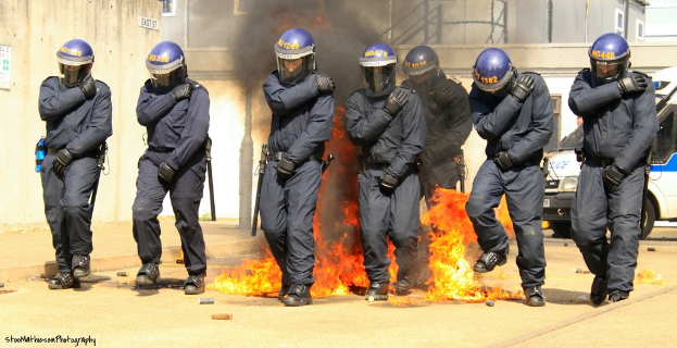 Menschen mit Helmen vor einem Feuer mit Gegenständen auf dem Boden, Gebäuden im Hintergrund, einem Fahrzeug, einem Plakat und einer Tafel an der linken Wand und Text unten.