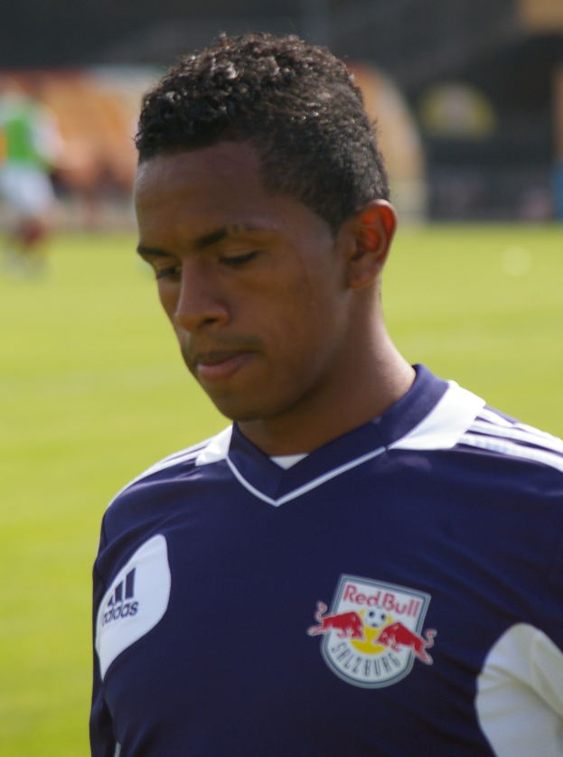 Ein junger Mann in einem blauen T-Shirt steht auf einem Fußballfeld, der als Flügelspieler mit kurzen dunklen Haaren und einem entschlossenen Gesichtsausdruck erscheint, vor einem unscharfen Hintergrund.