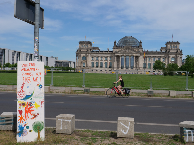 Eine Person in einem schwarzen Kleid fährt mit dem Fahrrad die Straße vor dem Reichstag in Berlin, Deutschland, entlang, mit Gebäuden, Bäumen und ein paar Menschen im Hintergrund unter einem blauen und weißen Himmel.