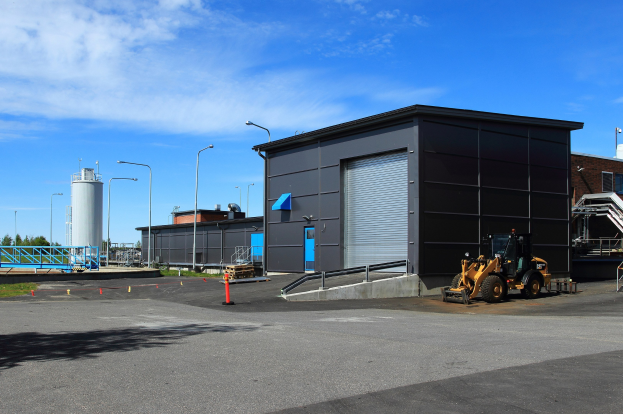 Großes Gebäude mit einem Bagger davor, umgeben von Straßeninfrastruktur und Fahrzeugen, unter einem bewölktem Himmel, als neue Industriepark-Anlage gekennzeichnet.
