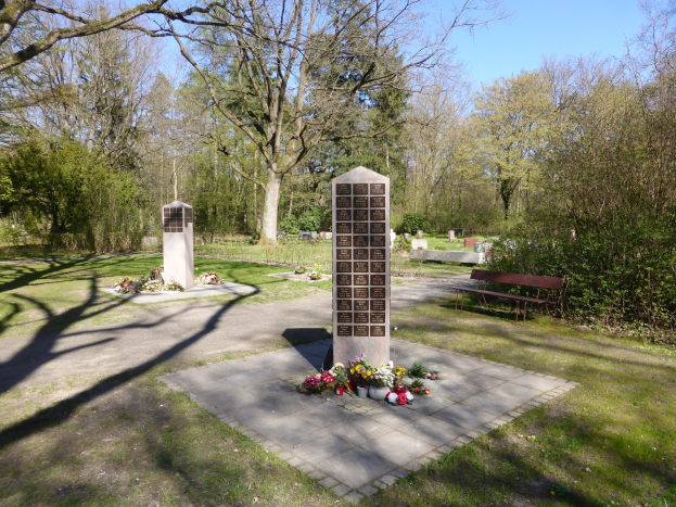 Kriegsdenkmal auf einer grünen Wiese mit Bäumen, Blumensträußen, einer Bank, Gräbern und einem klaren blauen Himmel.