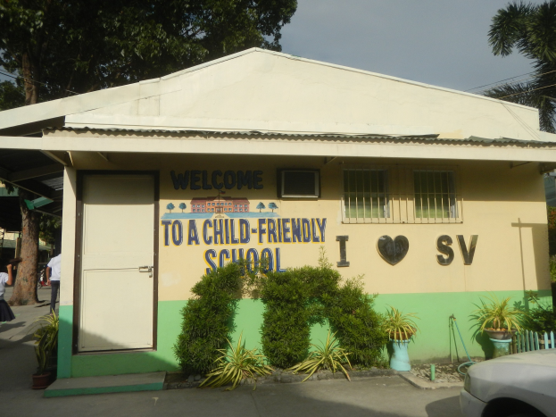 Ein einstöckiges Schulgebäude mit einem davor geparkten Auto, das eine Tür, Fenster und ein Schild "Willkommen in einer kinderfreundlichen Schule" zeigt, umgeben von Topfpflanzen, Bäumen, Menschen, Strommasten und Drähten unter einem bewölkten Himmel.