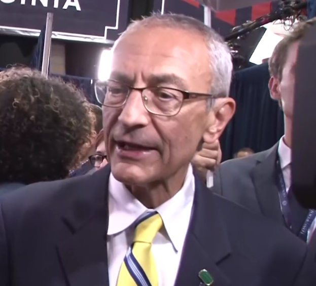 Ein Mann im Anzug und mit Krawatte, der eine Brille trägt, spricht vor Reportern mit einem Mikrofonständer vor sich, umgeben von einer Gruppe von Menschen, mit Scheinwerfern und einer Tafel im Hintergrund.