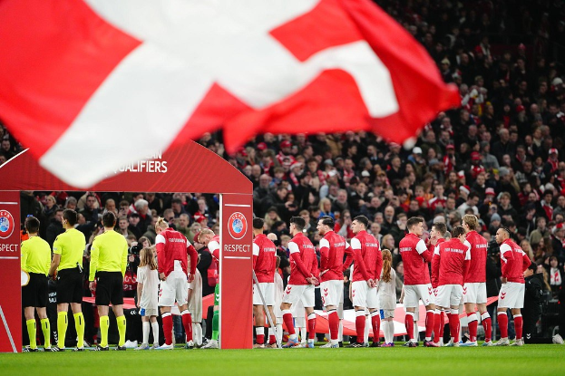 Eine Gruppe von Menschen auf einem Fussballfeld mit einer roten und weissen Flagge im Vordergrund, einem Bogen mit dem Text "Bayern Munich vs Bayern Munich Betting Tips & Preview" im Hintergrund und einer grossen Menge im Stadion.