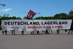 Eine Gruppe von Menschen, die ├Ąu├čerlich mit einem Banner stehen, auf dem "Deutschland, Lagerland Migration ist kein Verbrechen" und einer Flagge, mit Laternenmasten, Stra├čenlaternen, B├Ąumen, Kr├Ąnen, einem Geb├Ąude und einem bew├Âlktem Himmel im Hintergrund.