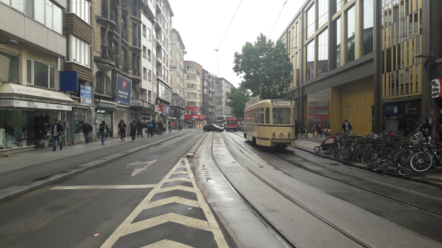 Eine Stadtstraße mit einer gelben Tram, Gebäuden mit Fenstern, Schildern, Fußgängern, parkenden Fahrrädern, einem Straßenpfahl, Drähten, einem Baum und einem bewölkten Himmel.