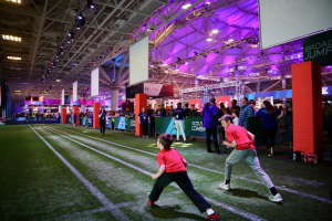 Zwei junge Jungs beim Fußballspielen auf einem Feld, umgeben von einer Gruppe von Menschen, mit Flaschen und anderen Gegenständen rechts, einem Zaun, Bannern, einem Anzeigebildschirm, Metallrahmen, einem Dach mit Deckenleuchten und Metallpfosten im Hintergrund.