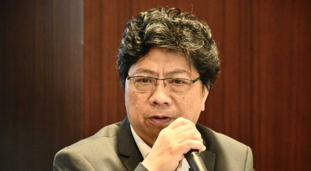 Ein Mann im Anzug mit Mikrofon, der lebhaft mit einer hölzernen Wand im Hintergrund spricht.