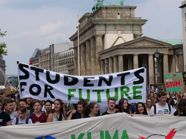 Gruppe von Schülern marschiert in Berlin mit einem leuchtend bunten "Students for Future"-Schild an Gebäuden, Bäumen und Himmel vorbei.