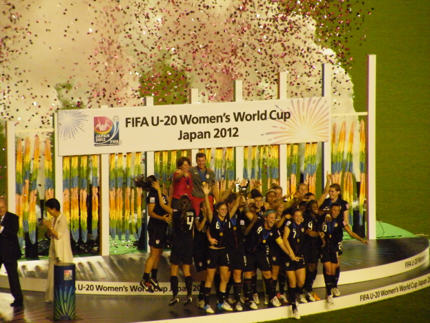 Eine Gruppe von Fussballspielerinnen in schwarzen Uniformen feiert ihren Sieg auf einer Bühne, umgeben von einer Menge mit Kameras, mit einem dekorierten Hintergrund und einem grasbedeckten Boden.