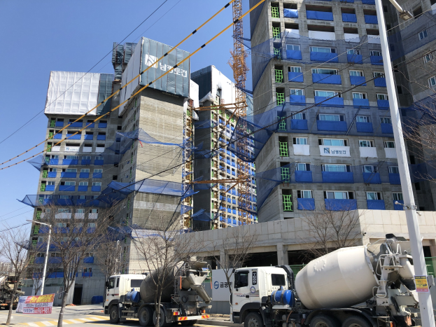 Baustelle mit einem Betonmischer-Lkw, der vor einem hohen Bauwerk im Bau parkt, Fahrzeugen auf der Straße, Bäumen, Strommasten, Holzplanken, Kabeln im Vordergrund und Himmel im Hintergrund.