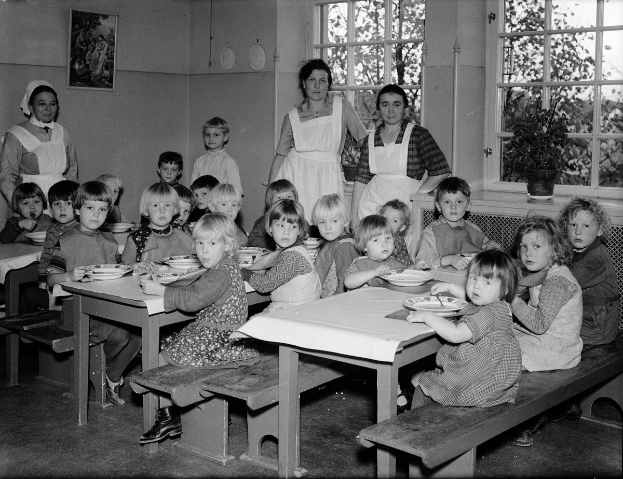 Kinder sitzen an Tischen in einem Klassenzimmer und essen Mahlzeiten, mit Bänken, einem Rahmenfoto an der Wand und Fenstern, die Pflanzen draußen zeigen.