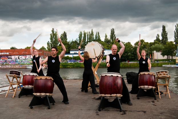 Fünf Menschen in schwarzen Kleidern spielen Musikinstrumente, darunter Trommeln, auf einem Stuhl, umgeben von Wasser, Bäumen und einem bewölkten Himmel, mit einem Gebäude im Hintergrund.