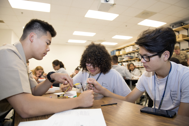 Drei Schüler an einem Tisch mit Papieren, Stiften, einem Handy und anderen Gegenständen, die an einem Projekt in einem Klassenzimmer mit Regalen, Deckenleuchten und einem Projektor im Hintergrund arbeiten.