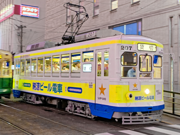 Eine gelbe und blaue Tram auf einer Stadtstraße bei Nacht mit Menschen drinnen, Gebäude mit Fenstern, Strommasten mit Drähten und Schilder im Hintergrund.