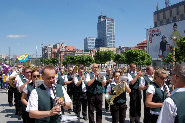 Eine Gruppe von Menschen in einheitlichen Kostümen, die verschiedene Musikinstrumente spielen, vor einem Hintergrund aus Bäumen, Pfählen, Fahnen und einem Banner mit Text und Bildern.