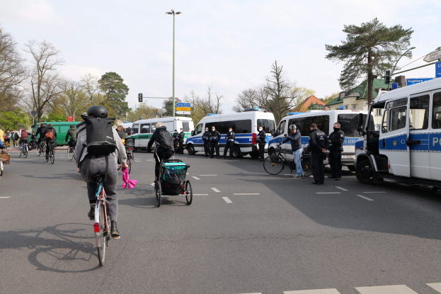 Eine Gruppe von Menschen, die Fahrräder auf einer Straße fahren, die von Polizeiwagen und anderen Fahrzeugen gesäumt ist, einige tragen Helme und Taschen, andere schieben Kinderwagen, mit Bäumen, Laternenmasten, Verkehrszeichen, Schildern, Gebäuden und einem bewölkten Himmel im Hintergrund.