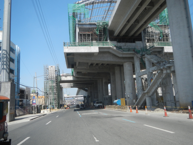 Eine vielbefahrene Straße mit fahrenden Fahrzeugen, Verkehrskegel an den Seiten, ein großes Gebäude im Hintergrund, sichtbarer Himmel über einer Brücke und Strommasten mit Drähten entlang der Straße.