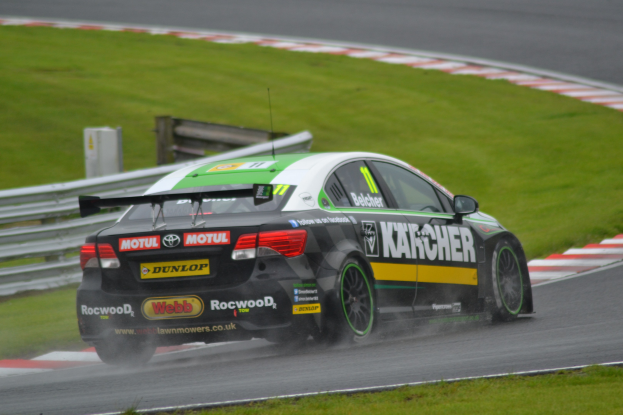 Ein Auto mit Text an der Seite, das auf einer regennassen Rennstrecke fährt, die von Gras und einer Holzkonstruktion umgeben ist.