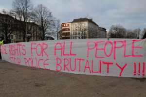 Eine Gruppe von Menschen hält ein Transparent mit der Aufschrift 'Rechte für alle Menschen Stoppt Polizeigewalt' auf dem Boden mit einer Straßenlaterne, einem Schild, Bäumen, Gebäuden mit Fenstern und einem bewölkten Himmel im Hintergrund.